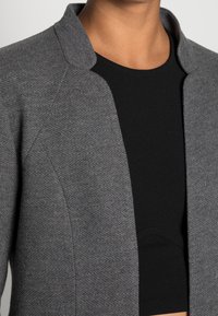 Šedý herringbone blazer se šálovým límcem, otevřeným předním dílem a texturovanou tkaninou, zkombinovaný s přiléhavým černým topem pod ním.