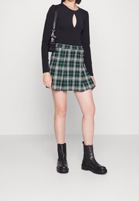 Schwarz langärmliges Oberteil mit einem Schlitz im Schlüsselbereich, kombiniert mit einem grünen karierter Faltenrock. Schwarze Stiefeletten runden das Outfit ab.