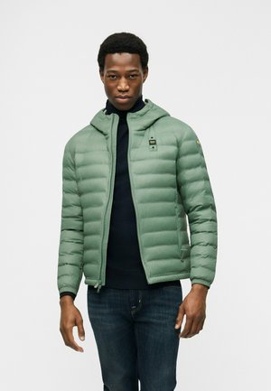 Giovane uomo che indossa una giacca trapuntata con cappuccio verde chiaro sopra un dolcevita nero e jeans blu scuro su sfondo semplice.