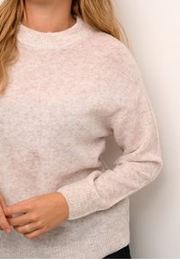 Frau, die einen hellrosafarbenen, strukturierten Strickpullover mit geripptem Kragen und Bündchen trägt, steht vor einer schlichten weißen Wand.