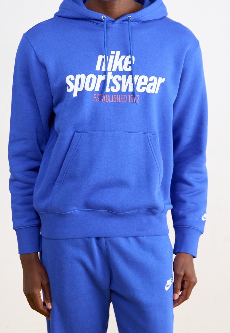 Blå Nike-hoodie med vit logotyp och text, blandmaterial av bomull, frontficka i kängurustil, ribbade cuffs och nederkant, avslappnad passform.