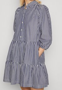 Robe à rayures bleues et blanches avec un devant boutonné, un col, des manches mi-longues et une jupe à volants. Comprend des poches et une coupe décontractée.