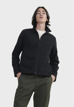 Schwarze Fleecejacke mit hohem Kragen, durchgehendem Reißverschluss und langen Ärmeln, kombiniert mit grünen Hosen. Glatte Textur und minimalistisches Design.