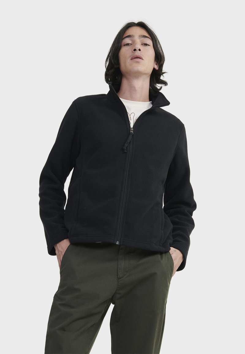 Schwarze Fleecejacke mit hohem Kragen, durchgehendem Reißverschluss und langen Ärmeln, kombiniert mit grünen Hosen. Glatte Textur und minimalistisches Design.