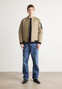 Parka em caqui, com um design acolchoado, punhos canelados e fecho de correr na frente. Combinado com jeans azuis e sapatos pretos.