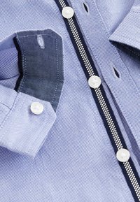 Chemise bleu clair texturée avec boutons blancs, bordure rayée bleu marine et blanc le long de la ligne de boutons, et poignet plié montrant la boutonnière.
