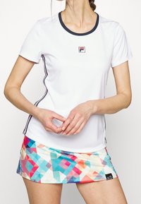 Bílé sportovní tričko s krátkým rukávem a černým výstřihem a bočními pruhy, které je kombinováno s barevně vzorovanou minisukní s geometrickými tvary.
