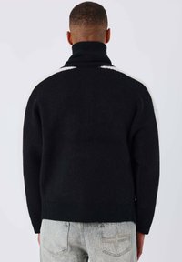 Homme ayant les cheveux courts, portant un pull à col roulé noir avec des rayures blanches sur les épaules et un jean délavé, de dos devant un fond uni.