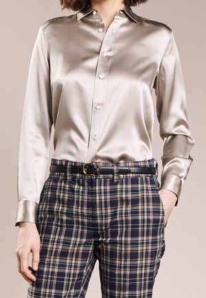 Femme portant une chemise beige satinée brillante boutonnée, rentrée dans un pantalon à carreaux bleu marine avec une ceinture noire, debout avec une main dans la poche.