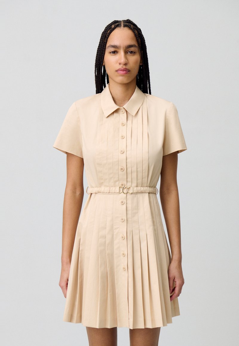 Vestido beige de manga corta con cuello de solapa, cierre de botones en la parte delantera, falda plisada y detalle de cintura fruncida con un anillo de metal.