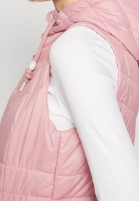 Ljusrosa pufferväst med huva, med kviltad textur, dragskojustering och öppna ärmhål, bärs över en vit ribbad topp.