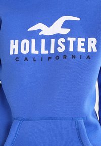 Blå huvtröja i mjukt material, med en stor vit "Hollister"-logotyp och mindre text "California". Inkluderar en framficka.
