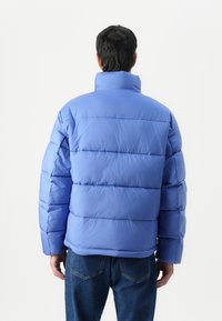 Lyseblå pufferjakke med høj krave, ribbede manchetter og vandret syninger. Båret over mørkeblå jeans. Glat, dunfyldt materiale.