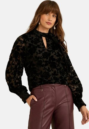 Juodas aksominis bluzonas su gėlių tekstūra, aukštu kaklu, skylių detalėmis ir pūstomis rankovėmis, derinamas su burgundinės spalvos dirbtinės odos kelnėmis.