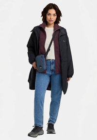 Schwarzer langer Mantel über einem dunkelroten Fleecepullover, weißem T-Shirt, blauer Jeanshose und dunklen Sneakers. Eine kleine marineblaue Umhängetasche wird getragen.