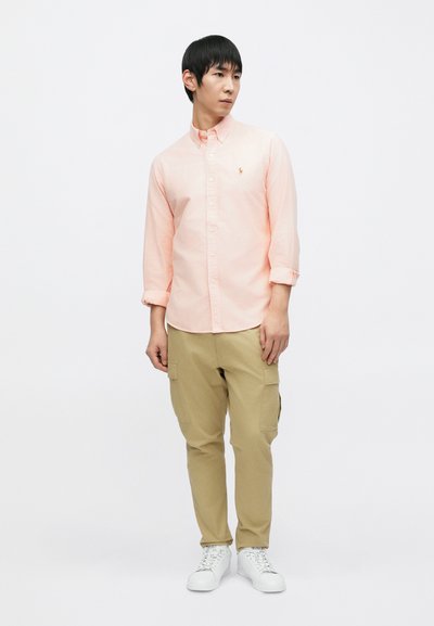 Ανοιχτό ροζ πουκάμισο με κουμπιά και μακριά μανίκια, σε συνδυασμό με μπεζ παντελόνι cargo. Το μοντέλο φοράει λευκά αθλητικά παπούτσια, στέκεται σε απλό φόντο.