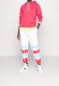 Felpa rossa con tasca frontale e lacci bianchi, indossata sopra una maglietta gialla; pantaloni jogger bianchi con strisce tie-dye rosse e blu; sneakers bianche.