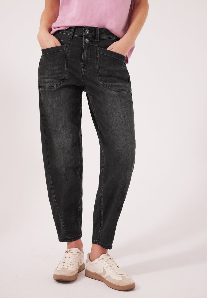 Personne portant un jean noir délavé avec de grandes poches avant, une chemise rose et des baskets blanches, debout les mains dans les poches.