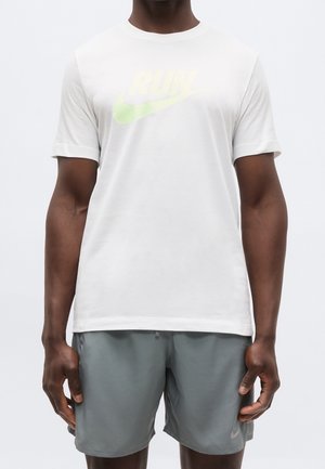 Mann trägt ein weißes Nike "RUN" T-Shirt und graue Nike Sportshorts, steht mit entspannten Armen an den Seiten vor einem einfarbigen Hintergrund.
