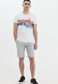 T-shirt blanche avec un graphisme abstrait de vagues multicolores, associée à un short gris clair et des baskets noires. Design simple et décontracté.