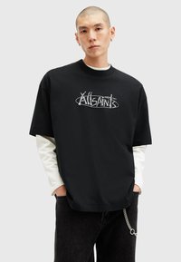 Sort oversized t-shirt med hvidt grafisk logo, korte ærmer, rund hals, lagdelt med en hvid skjorte med lange ærmer nedenunder, parret med mørke jeans.