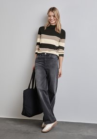 Gestreifter Pullover in Dunkelgrün, Beige und Creme; kombiniert mit dunkelgrauen, weiten Jeans. Hält eine schwarze, gesteppte Tragetasche; weiße Sneaker.