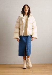 Doudoune blanche avec un design court, pull crème à côtes, jupe en denim longueur genou et bottes beiges à lacets sur un sol en bois.