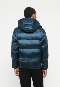 Persona que lleva una chaqueta acolchada de color verde azulado oscuro con capucha, de espaldas, contra un fondo blanco liso.