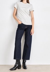 Grå bomullströja med korta ärmar och textdesign, parat med mörkblå denimjeans. Modellen har på sig svarta ankelboots med klack.