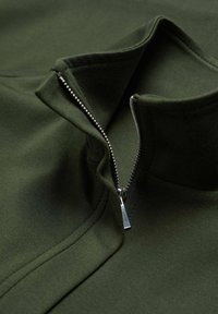 Tela texturizada de color verde oscuro con un cuello alto y cierre de cremallera. Herrajes de cremallera plateados con una lengüeta. Diseño liso y ajustado.