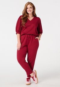 Bordeaux jumpsuit met een V-hals, korte pofmouwen en een koordtaille. Gecombineerd met roze sneakers met rode accenten. Soepele textuur.