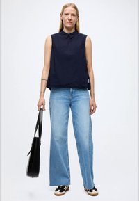 Top senza maniche blu navy con colletto, abbinato a jeans a gamba larga azzurri. Borsa nera in mano, sneakers basse con dettagli crema e neri.