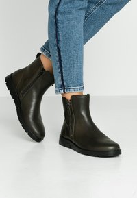 Bottes basses en cuir vert foncé avec une fermeture éclair latérale, un bout rond et une semelle en caoutchouc noir texturée. Portées avec un jean bleu raccourci.