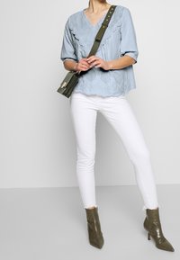 Světle modrá blůza s krajkovými detaily, výstřihem do V a krátkými rukávy, v kombinaci s bílými skinny džíny a olivově zelenými kotníkovými botami.