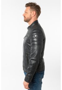 Schwarze Lederjacke mit gesteppten Schulterakzenten, seitlicher Reißverschlusstasche und Stehkragen, seitlich an einem Modell in blauen Jeans gezeigt.