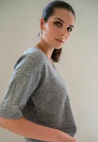 Femme avec des cheveux bruns attachés, portant des boucles d'oreilles en or et un haut en maille gris, regardant par-dessus son épaule sur un fond uni.