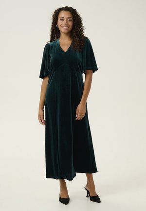 Long, deep green velvet dress with a V-neck, short wide sleeves, and a relaxed fit, complemented by black high-heeled shoes.