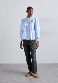 Lys blå bomullsskjorte med broderi og smocking-detaljer, lange ermer, rund halslinning, kombinert med mørke jeans og brune slip-on sko.