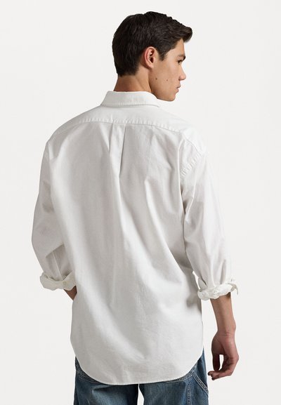 Camisa de algodón blanca con cuello extendido, mangas arremangadas y un pliegue en la parte posterior. Corte ajustado con un dobladillo redondeado y textura suave.