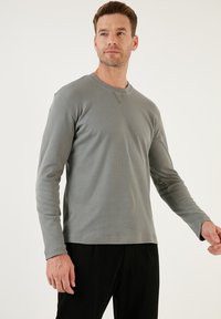 Szürke, hosszú ujjú póló ribbed anyagból, környakú kialakítással és diszkrét varrással a galléron. Fekete nadrággal viselve.