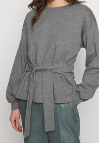 Pullover grigio a maniche lunghe con vita aderente e dettaglio della cintura legata. Tessuto morbido, leggero design a peplo con polsini a coste.