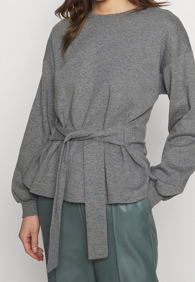 Pullover grigio a maniche lunghe con vita aderente e dettaglio della cintura legata. Tessuto morbido, leggero design a peplo con polsini a coste.
