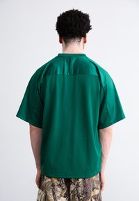Zöld, rövid ujjú póló hálós textúrával, sima, egyszínű résszel a tetején. Mintás camouflaged sorttal viselve.