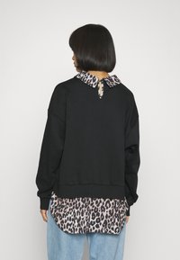 Kvinna med rakt svart hår bär en svart tröja över en blus med leopardmönster och ljusblå jeans, vänd bort mot en enkel bakgrund.