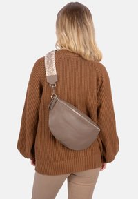 Braune Leder-Umhängetasche mit geschwungener Form, ausgestattet mit einem texturierten Riemen im geometrischen Muster. Der Reißverschluss ist an der Tasche sichtbar.