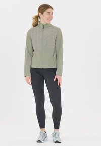 Ljusgrön idrottsjacka med en kviltad grå bröstpanel, dragkedja fram och två sidofickor. Matchas med svarta leggings och grå sneakers.