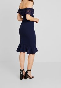 Marinblå, off-the-shoulder klänning med spetsdetaljer och volangnederdel. Fittad silhuett med utställd nederdel. Svarta klacksandaler.