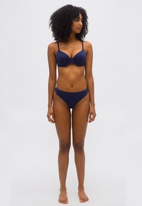 Femme aux cheveux bouclés debout pieds nus, portant un soutien-gorge bleu marine et une culotte assortie, regardant vers sa droite sur un fond uni.