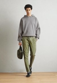 Hanorac gri cu glugă, pantaloni tapered verde măsliniu, adidași negri, ținând o geantă închisă la culoare. Material neted, croială lejeră, design minimalist.