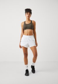Atletisk sports-bh i olivengrønn med en glatt tekstur, sammen med hvite shorts med elastisk midje og svarte sidelinjer.